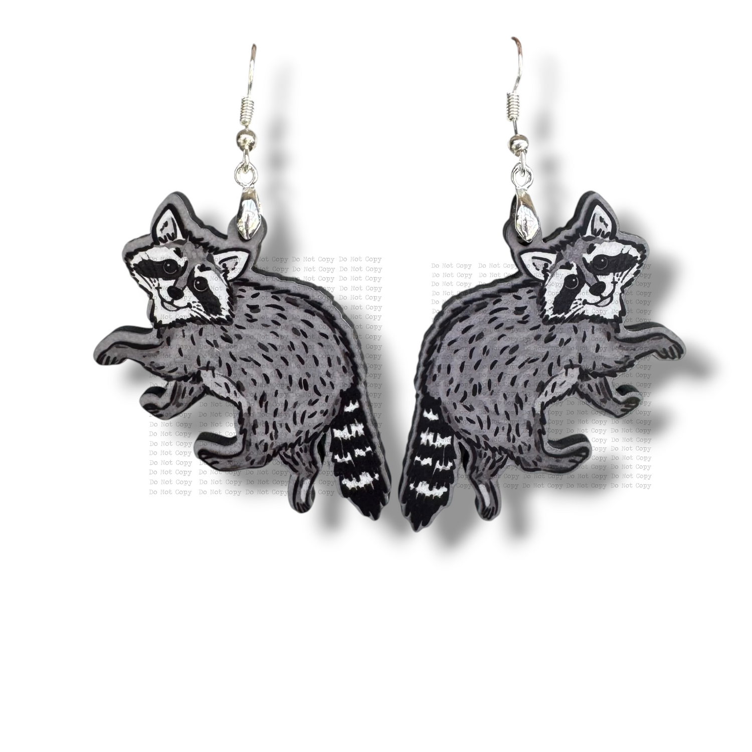 Two illustrated raccoon earrings made of gray and black MDF hanging from silver hooks. The raccoons are shown in a playful crawling pose with detailed fur textures and striped tails, set against a clean white background.