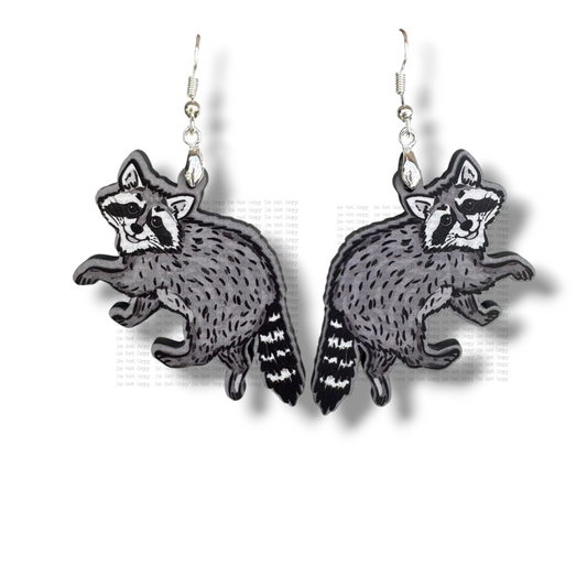 Two illustrated raccoon earrings made of gray and black MDF hanging from silver hooks. The raccoons are shown in a playful crawling pose with detailed fur textures and striped tails, set against a clean white background.