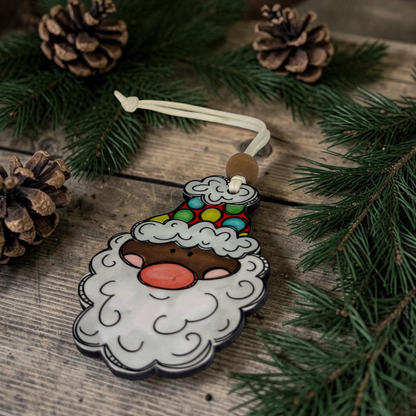 A Santa Claus-shaped Christmas ornament with a white beard, colorful hat, and cream hanging cord, photographed on a rustic wooden surface with pine branches and pinecones around it.