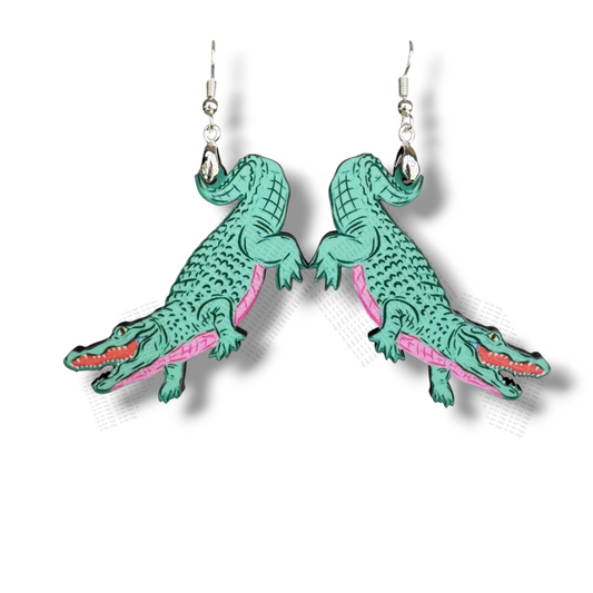 A pair of dangle earrings featuring teal-colored alligators with bright pink underbellies. The alligators are illustrated in a detailed, cartoon style with open mouths, hanging from silver hooks against a white background.