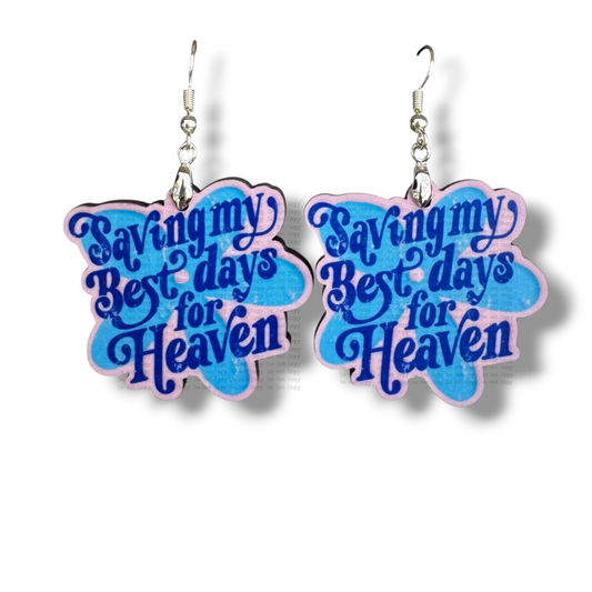 Two cloud-shaped MDF earrings featuring the text "Saving my Best days for Heaven" in a blue retro font. The design has a layered blue and light pink/lilac background with a dark purple outline, hanging from silver hooks on a white background