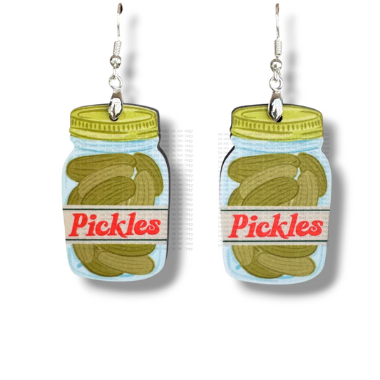 Two flat, illustrative earrings shaped like clear mason jars filled with green pickles. The jars have yellow lids and a white label with the word "Pickles" written in a retro red font, hanging from silver hooks against a white background.