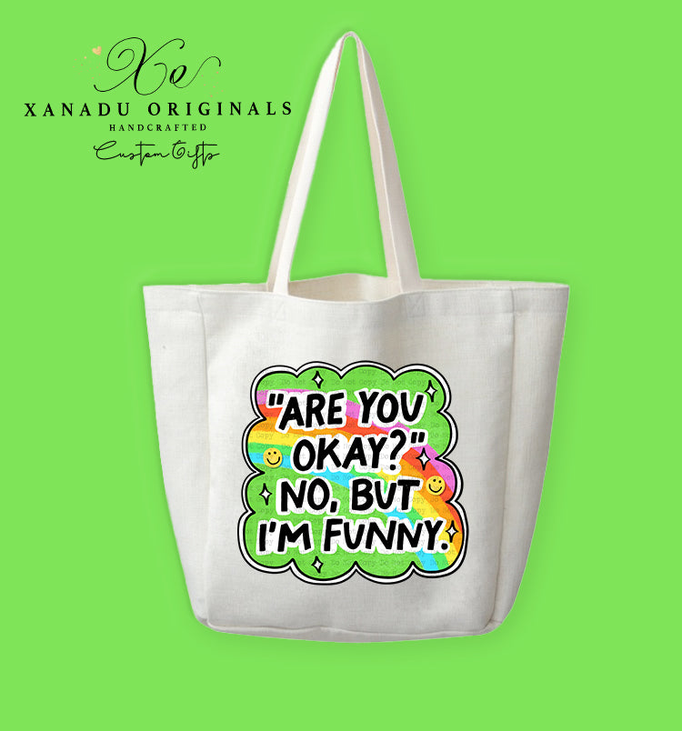 A cream-colored polyester linen tote bag featuring a vibrant retro cartoon graphic shaped like a wavy cloud outline. Inside the cloud is a bright rainbow pattern, yellow smiley faces, and white sparkle stars surrounding the bold black text that reads "'ARE YOU OKAY?' NO, BUT I'M FUNNY."