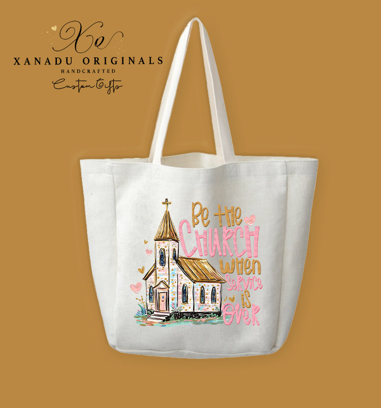 A cream-colored polyester linen tote bag featuring a watercolor sketch illustration of a brown and white country church with a cross steeple and floating pink hearts, paired with stylized pink and gold text that reads "Be the CHURCH when service is OVER."
