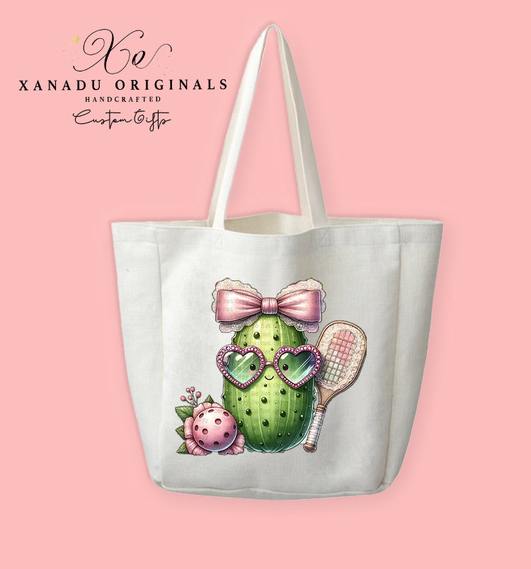 A cream-colored polyester linen tote bag featuring a cute illustration of a smiling pickle character wearing heart glasses and a big pink lace bow while holding a pickleball paddle.