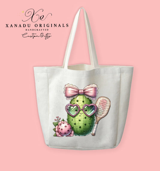A cream-colored polyester linen tote bag featuring a cute illustration of a smiling pickle character wearing heart glasses and a big pink lace bow while holding a pickleball paddle.