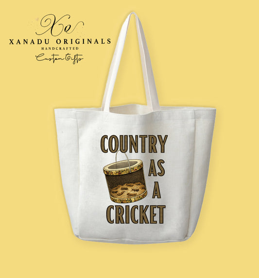 A cream-colored polyester linen tote bag featuring a watercolor illustration of a cylindrical metal wire cricket bait box with a camouflage lid and base, surrounded by text that reads "COUNTRY AS A CRICKET" in gold block lettering with dark outlines.
