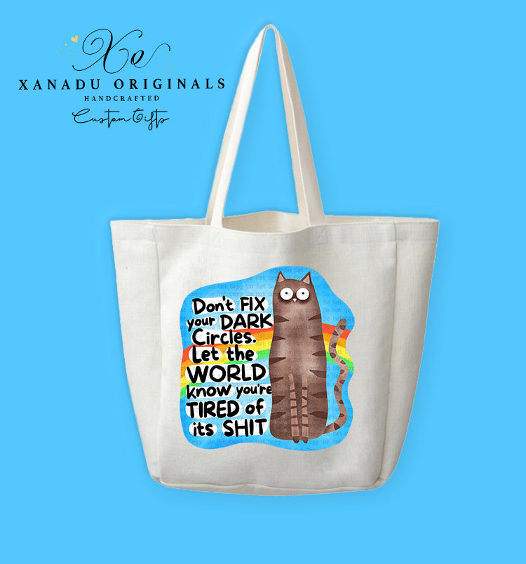 A cream-colored polyester linen tote bag featuring a cartoon illustration of a tall, wide-eyed brown tabby cat sitting in front of a retro rainbow within a blue cloud shape. To the left, stylized text reads "Don't FIX your DARK Circles. Let the WORLD know you're TIRED of its SHIT."