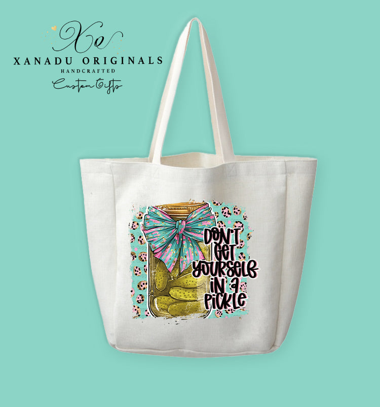 A cream-colored polyester linen tote bag featuring a vibrant leopard print graphic of a pickle jar tied with a teal and pink bow next to the text "DON'T GET YOURSELF IN A PICKLE".