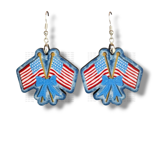 Two contoured MDF earrings shaped like two crossed American flags tied together with a blue bow. The design has a dark blue denim-textured border with scalloped edges and hangs from silver ear hooks on a white background
