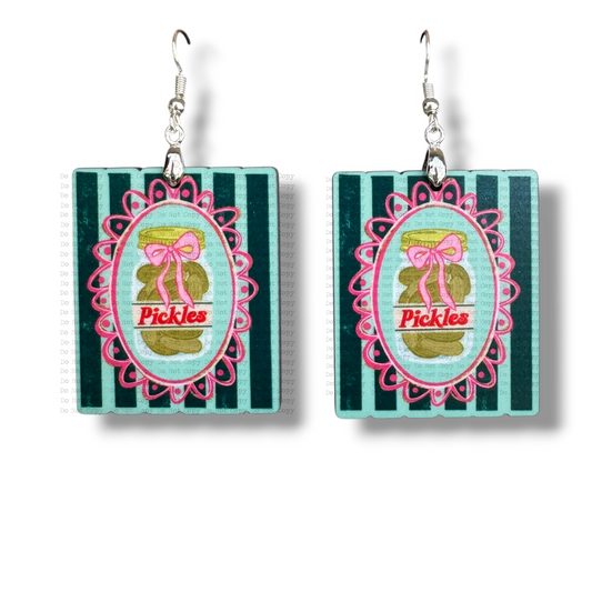 Square MDF earrings featuring a vintage-style pickle jar illustration with a pink bow. The jar is centered in a pink scalloped frame over a teal and dark green vertical striped background, hanging from silver hooks on a white background.