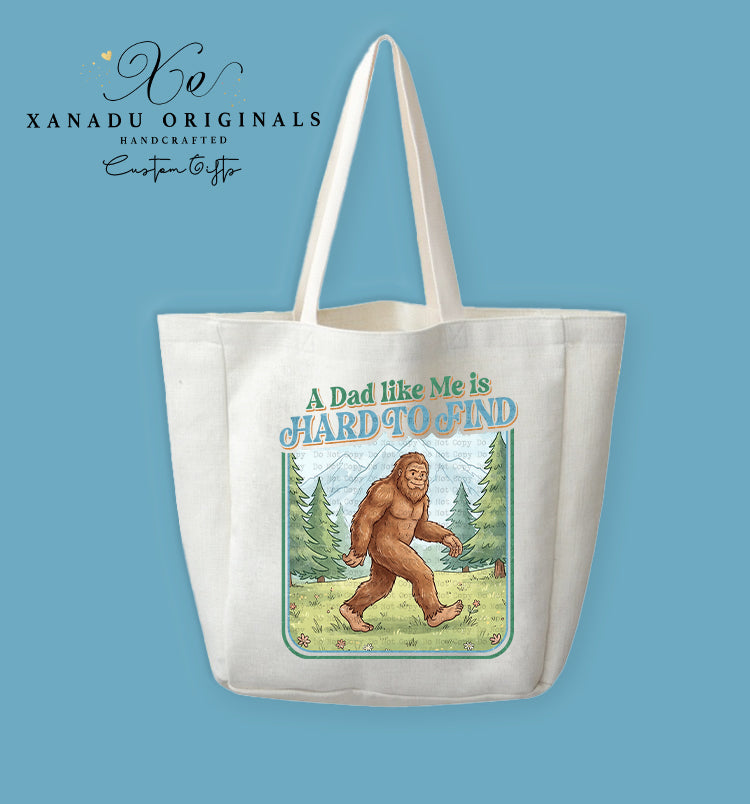 A cream-colored polyester linen tote bag featuring a retro cartoon illustration of a brown Bigfoot walking through a green pine forest with mountains under a light blue sky, framed by a thin green border, with the text "A Dad like Me is HARD TO FIND" written in stylized blue and green lettering above.