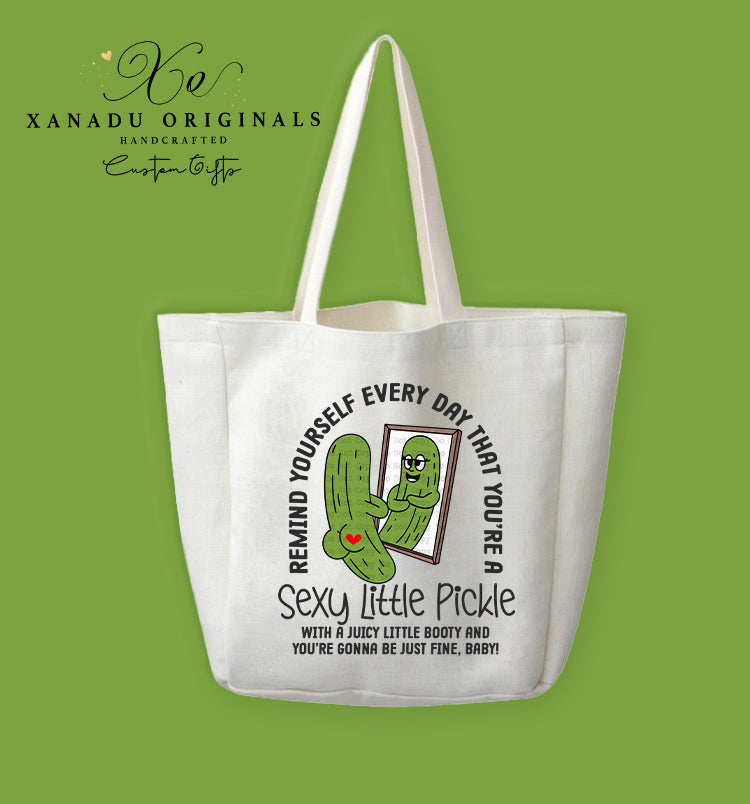 A cream-colored polyester linen tote bag featuring a funny cartoon illustration of a pickle looking in a mirror next to a motivational text overlay about being a sexy little pickle.