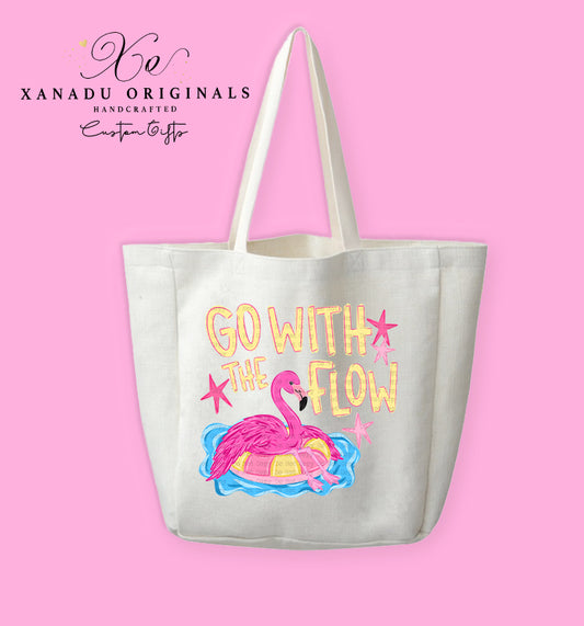 A cream-colored polyester linen tote bag featuring a cartoon illustration of a pink flamingo relaxing on a yellow patterned inner tube float in blue water, under vibrant yellow and pink text that reads "GO WITH THE FLOW" with small pink stars.