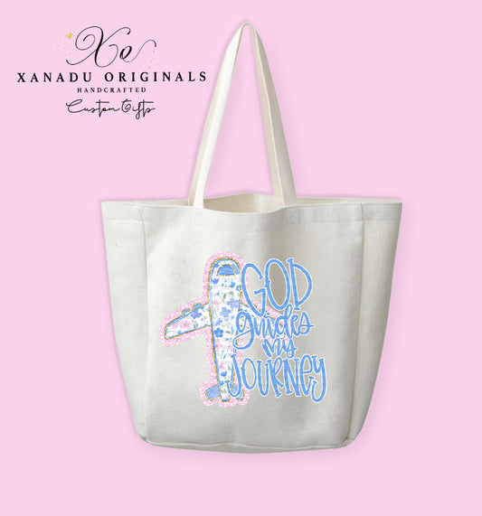 A cream-colored polyester linen tote bag featuring a cartoon airplane graphic covered in a blue floral pattern with a pink stitched border, paired with stylized blue bubble lettering that reads "GOD Guides My JOURNEY."