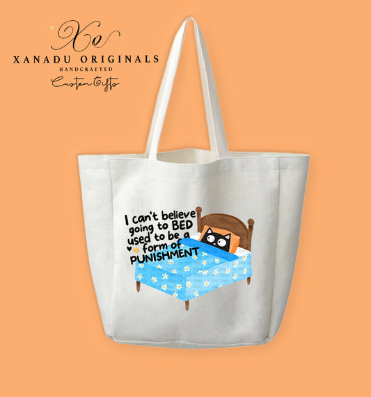 A cream-colored polyester linen tote bag featuring a cartoon illustration of a wide-eyed black cat in a wooden bed under a blue blanket with small white flowers. Black text to the left reads "I can't believe going to BED used to be a form of PUNISHMENT."
