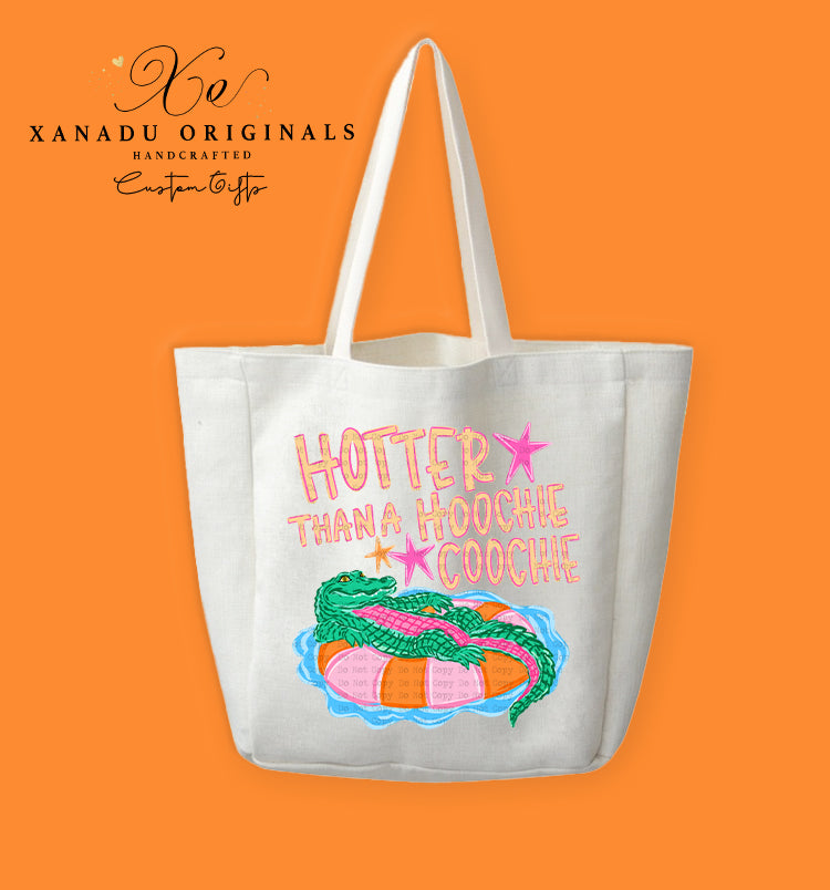 A cream-colored polyester linen tote bag featuring a cartoon illustration of a green alligator relaxing on a pink and orange striped inner tube float in blue water, under bright pink and yellow text that reads "HOTTER THANA HOOCHIE COOCHIE" with small pink stars.