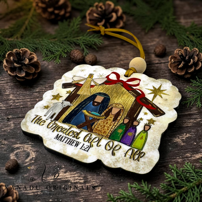 A decorative Christmas ornament shaped like a rustic manger scene, laying on a dark wooden surface with pine branches, pinecones, and small brown beads around it. The ornament shows Mary, Joseph, baby Jesus, shepherds, animals, stars, and the text 'The Greatest Gift Of All MATTHEW 1:21'.