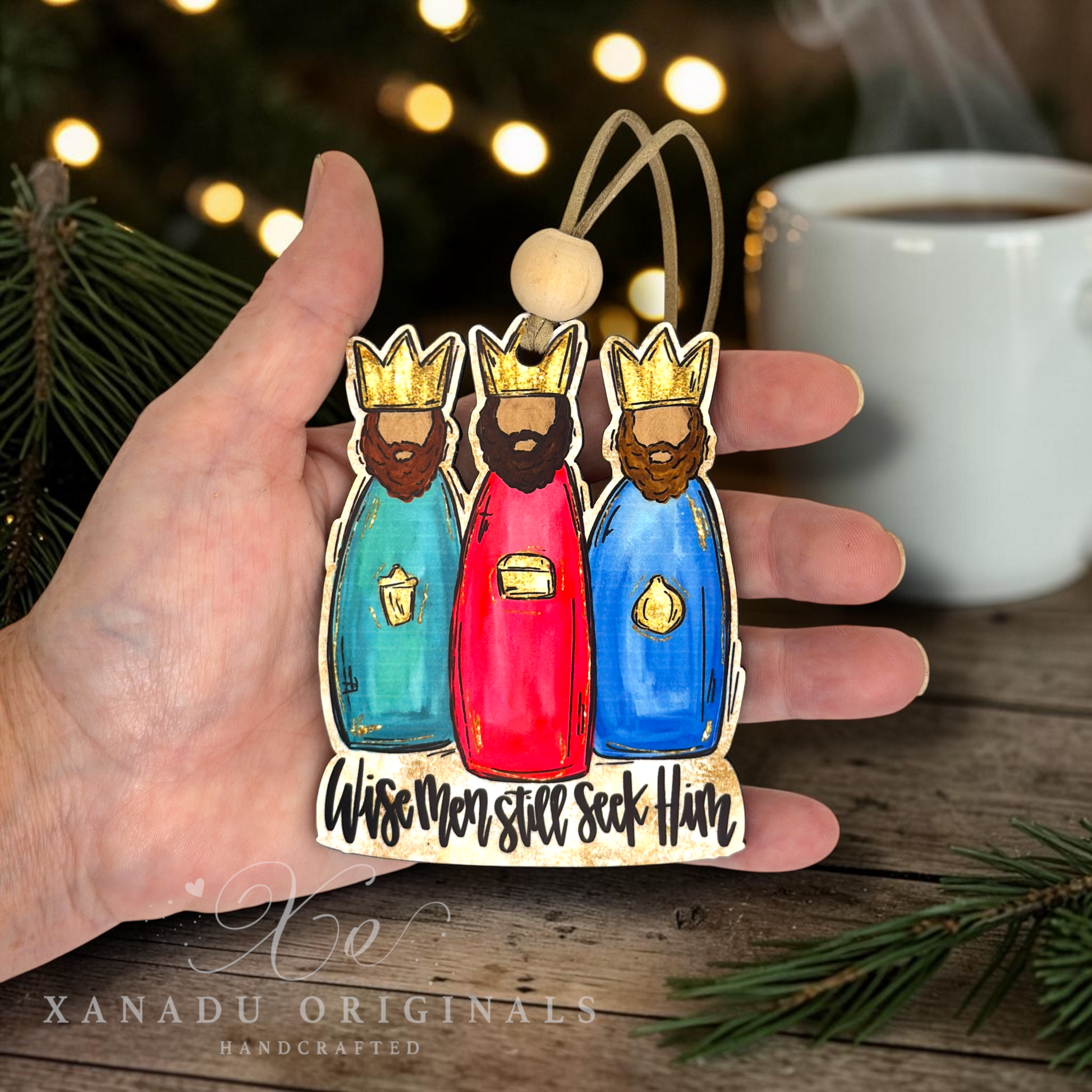 A hand holds a handcrafted wooden Christmas ornament shaped like three wise men in colorful robes with gold crowns and the text 'Wisemen still seek Him.' The background shows a blurred holiday scene with pine branches, warm lights, and a cup of coffee.