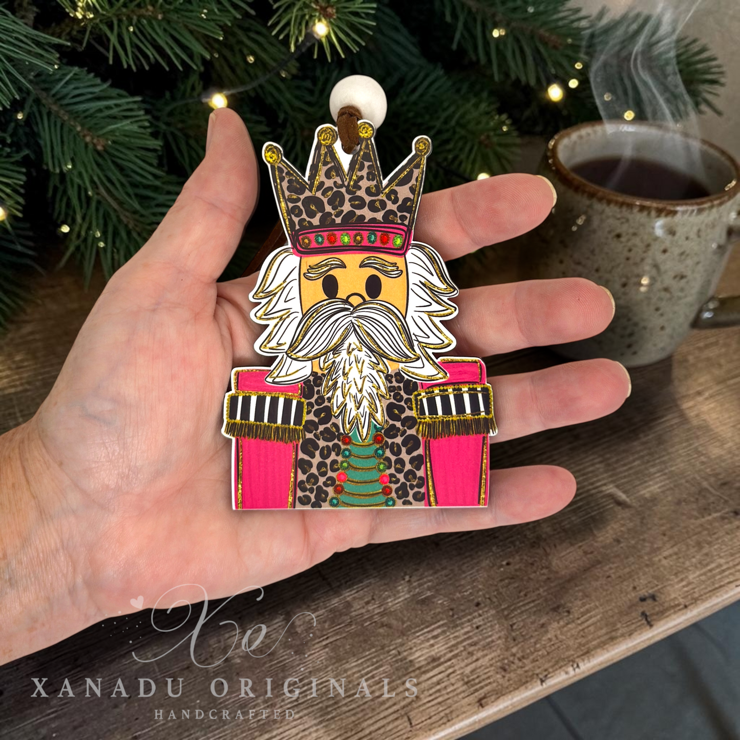 A hand holds a handcrafted wooden nutcracker ornament featuring a leopard-print uniform, white beard, and crown, with a Christmas tree and a mug of tea in the background.