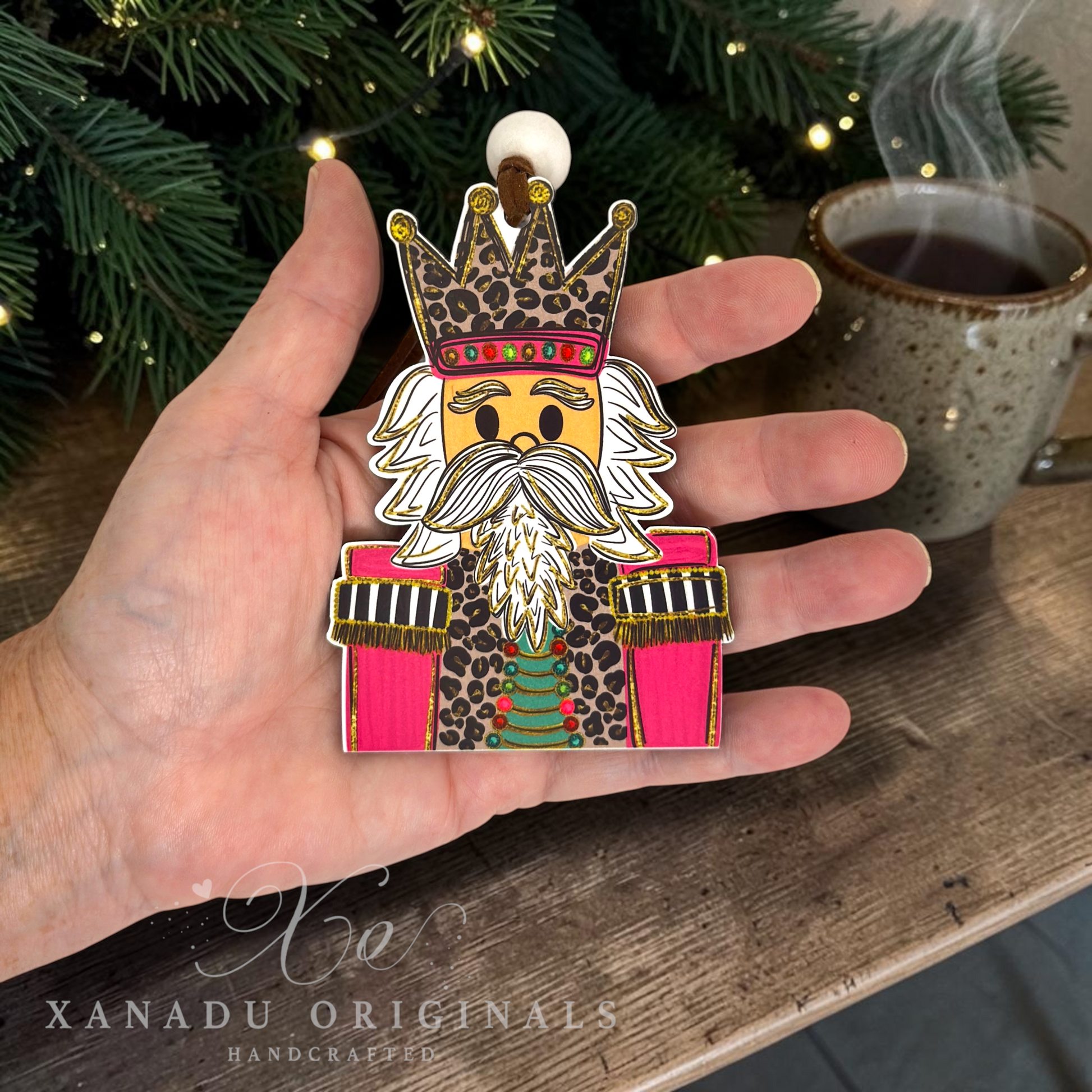 A hand holds a handcrafted wooden nutcracker ornament featuring a leopard-print uniform, white beard, and crown, with a Christmas tree and a mug of tea in the background.