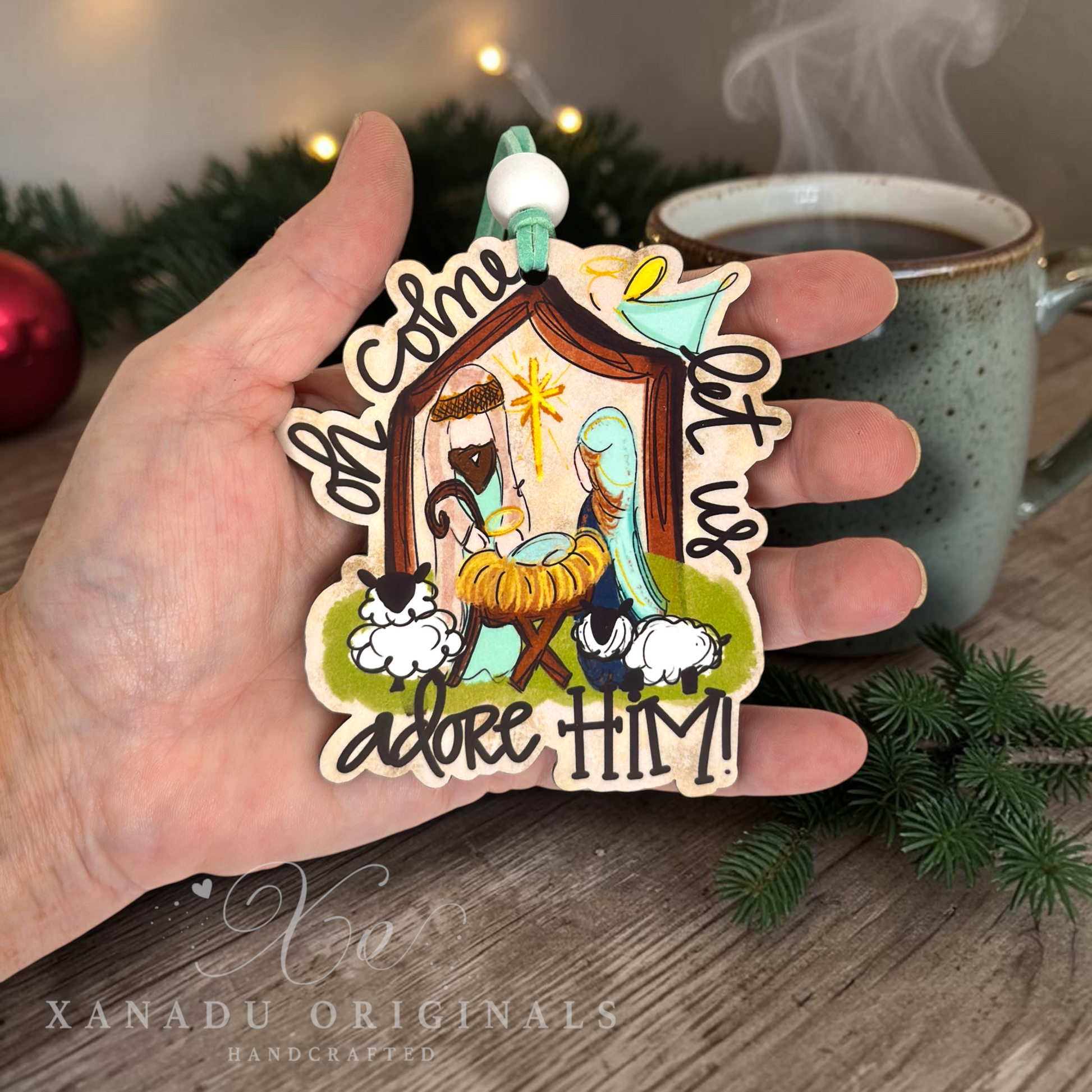 A hand holds a handcrafted wooden Christmas ornament featuring a nativity scene with the text 'Oh Come Let Us Adore HIM!'. The background shows a cozy holiday setting with a mug of coffee, pine branches, and warm string lights.