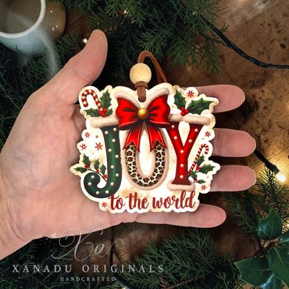A hand holds a handcrafted Christmas ornament spelling 'JOY' with holly, candy canes, and the phrase 'to the world.' The ornament has a cream base with red, green, brown, and leopard-print details, and a wooden loop at the top.