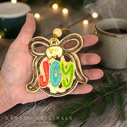 A hand holds a gold-plated Christmas ornament shaped like a bauble with a bow and the word 'Joy' in colorful red, blue, green, and yellow lettering. The background shows a cozy holiday scene with a cup of tea, warm string lights, and pine branches.