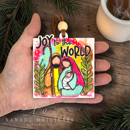 A hand holds a square wooden ornament featuring a colorful hand-painted nativity scene with the text 'Joy to the World.' The background includes a wooden surface, evergreen branches, and a small white cup.