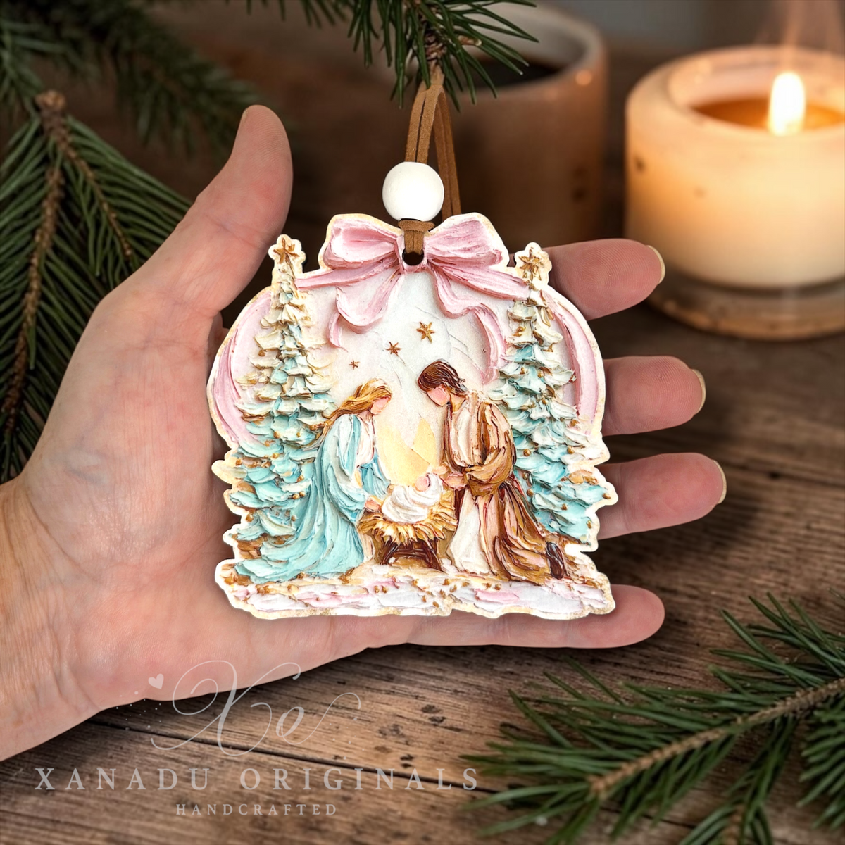 A hand holds a handcrafted Christmas ornament featuring the Holy Family, with a pink bow, gold accents, and green trees, set against a rustic wooden background with a burning candle.