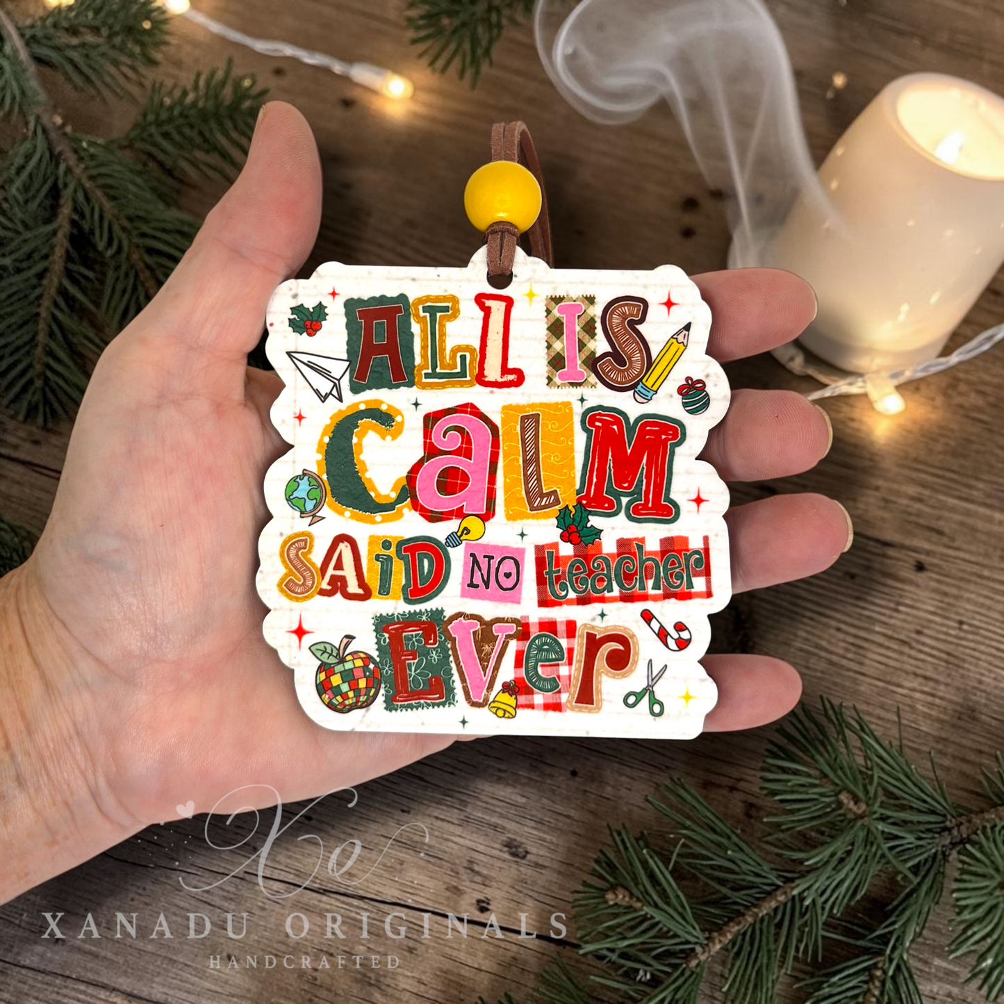 A hand holds a die-cut acrylic ornament featuring the phrase 'ALL IS CALM SAID NO TEACHER EVER' in colorful retro lettering with holiday-themed doodles. The background shows a rustic wooden surface with pine branches and warm string lights.