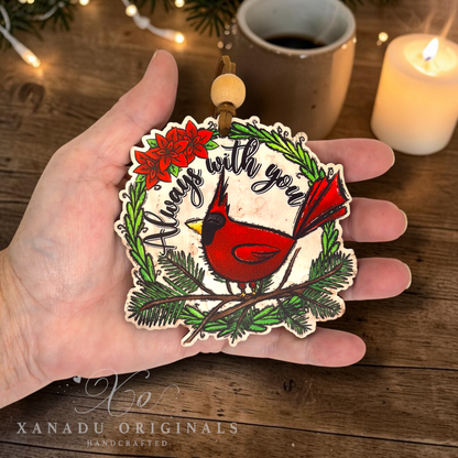 A hand holds a handcrafted wooden Christmas ornament featuring a red cardinal on a green pine wreath with the text 'Always with you'. The background shows a cozy holiday scene with a mug of coffee, a lit candle, and warm fairy lights.