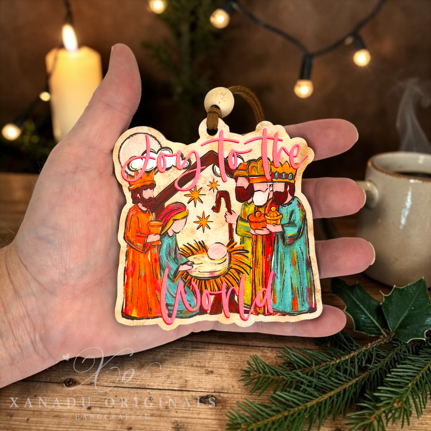 A hand holds a die-cut wooden Christmas ornament featuring a nativity scene with the text 'Joy to the World' in pink script. The background shows a cozy holiday setting with a lit candle, warm string lights, a mug, and pine branches.