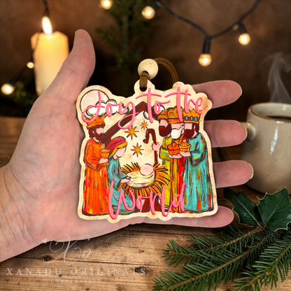 A hand holds a die-cut wooden Christmas ornament featuring a nativity scene with the text 'Joy to the World' in pink script. The background shows a cozy holiday setting with a lit candle, warm string lights, a mug, and pine branches.