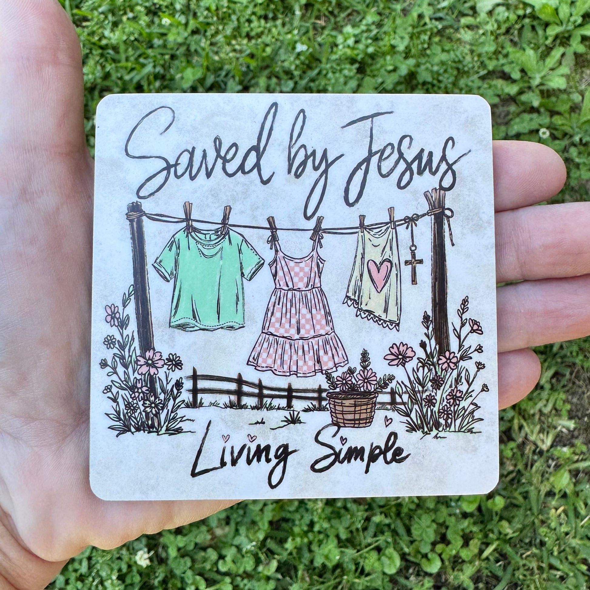 3.6 inch square MDF wood coaster with rounded corners featuring a sketch-style illustration of a clothesline with a green shirt, checkered pink dress, and heart tank top hanging next to a wooden cross, surrounded by wildflowers and text reading Saved by Jesus Living Simple.
