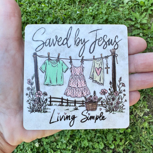 3.6 inch square MDF wood coaster with rounded corners featuring a sketch-style illustration of a clothesline with a green shirt, checkered pink dress, and heart tank top hanging next to a wooden cross, surrounded by wildflowers and text reading Saved by Jesus Living Simple.