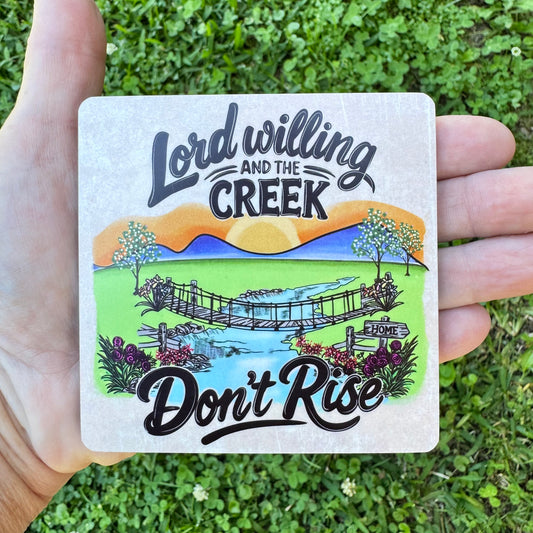 3.6 inch square MDF wood coaster with rounded corners featuring a scenic country landscape illustration with a footbridge over a creek, trees, flowers, a mountain sunset, and stylized text reading Lord Willing and the Creek Don't Rise on a textured background.