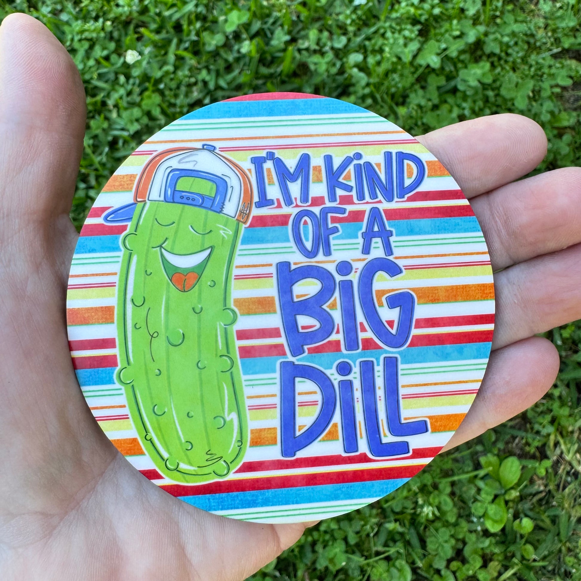 3.6 inch round circular MDF wood coaster featuring an illustration of a laughing cartoon green pickle wearing a blue and orange backward baseball cap with text reading I'm Kind of a Big Dill on a red, blue, green, and orange horizontally striped background.