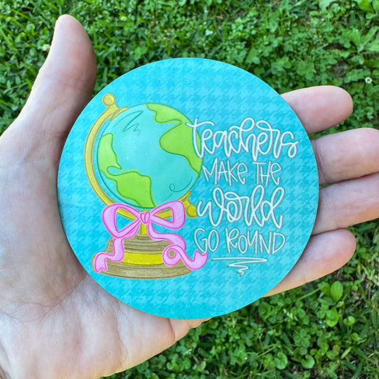 3.6 inch round circular MDF wood coaster featuring an illustration of a vintage green and yellow desk globe with a pink bow, stylized text reading Teachers Make The World Go Round on a turquoise houndstooth background.