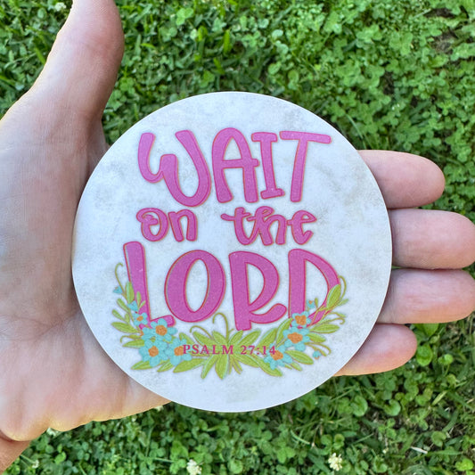 3.6 inch round circular MDF wood coaster featuring bold pink typography that reads Wait on the Lord with a small blue wildflower and green leaf garland below framing the text Psalm 27:14, all on a vintage rustic cream background.