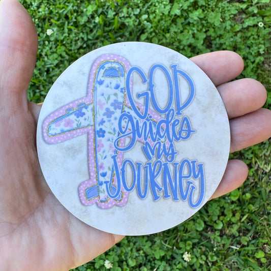 3.6 inch round circular MDF wood coaster featuring a stylized passenger airplane illustration patterned with blue flowers and pink polka dots, next to periwinkle blue typography reading God guides my journey on a rustic cream background.