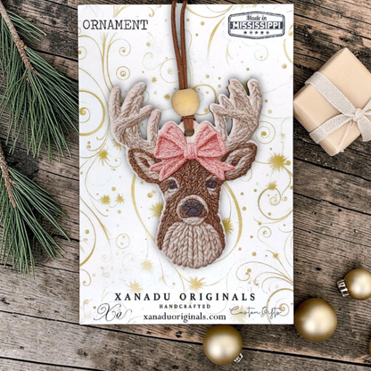 A product layout shot of a woodland deer wood ornament with a realistic knitted yarn pattern and light pink bow, mounted flat on a white Xanadu Originals presentation card.