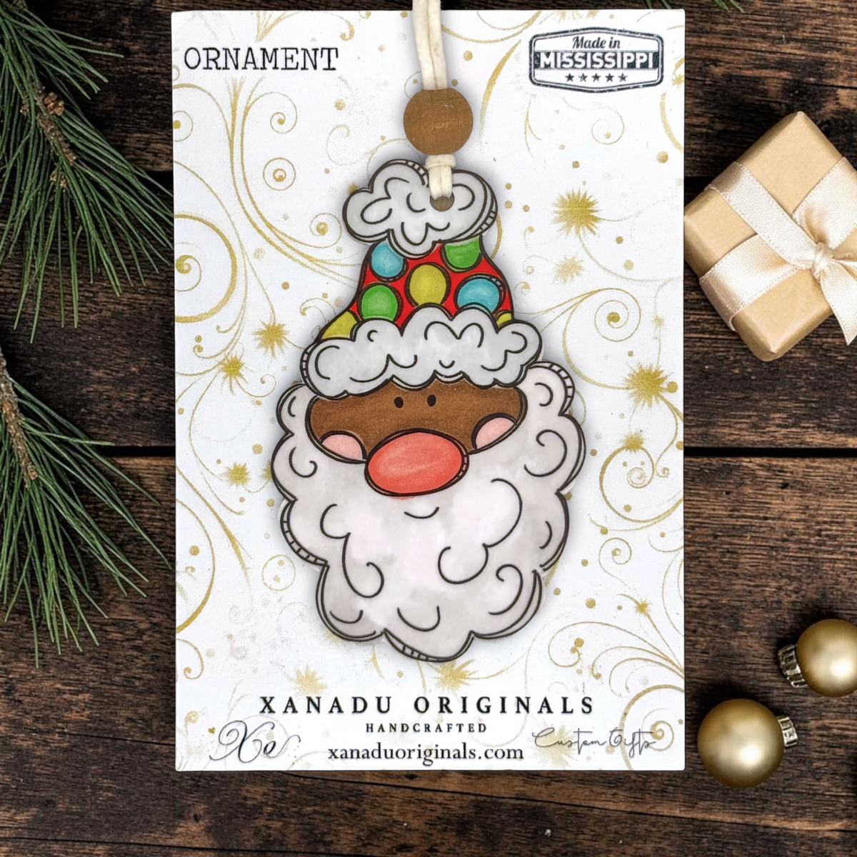 A product display card for a dark skin tone Santa wood ornament with a colorful polka dot hat, brown wood bead, and white cord hanger, sitting on a dark rustic wooden background with a gold gift box and pine needles.