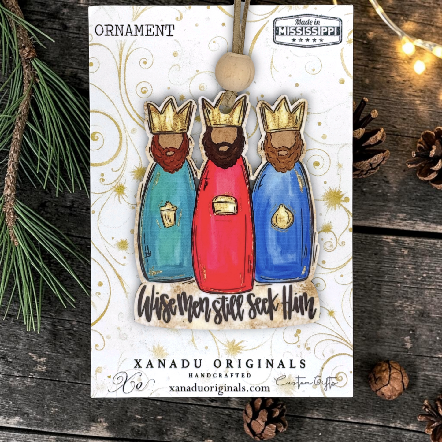 A handmade Christmas ornament featuring three crowned wise men in teal, red, and blue robes with gold accents, attached to a white card with gold swirls. The card is photographed on a rustic dark wood surface with pine branches and pinecones, and the ornament hangs from a wooden loop.