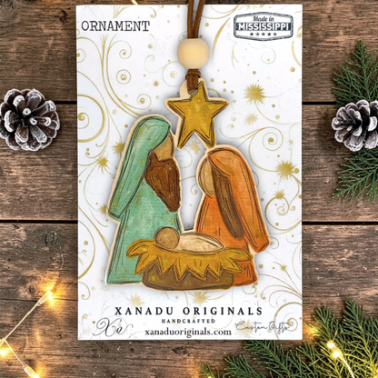 Handcrafted Christmas nativity ornament featuring Mary, Joseph, baby Jesus, and a gold star, displayed on its branded card over a rustic wooden background with pinecones and fairy lights.