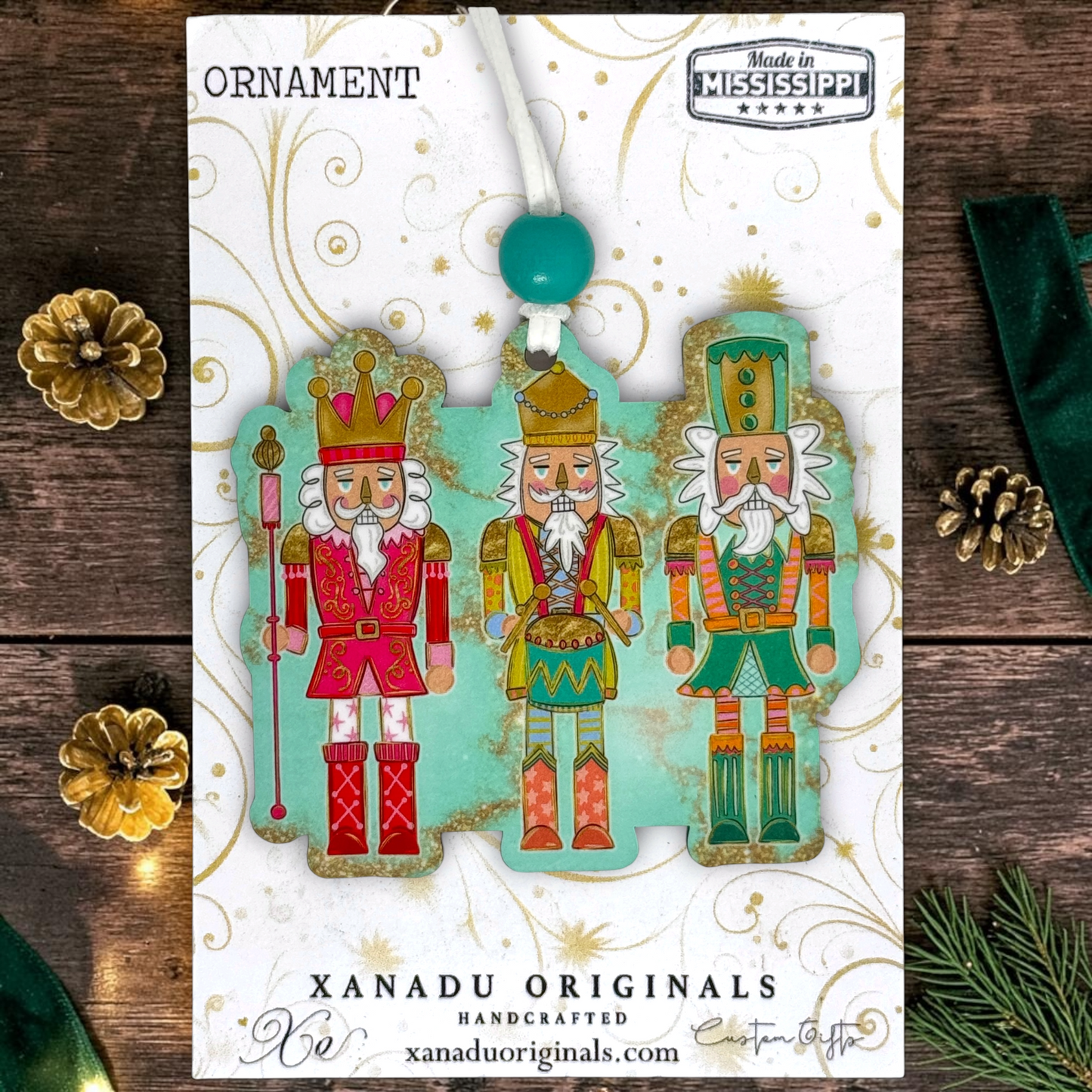A Christmas ornament package from Xanadu Originals featuring three colorful nutcracker figures on a white card with gold swirls. The package is photographed on a dark wood surface with decorative pine branches, pinecones, and a green ribbon.