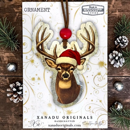 A custom-cut wood ornament showing a deer head in a red Santa hat with a vintage text background, mounted on a gold swirl display card next to pine sprigs and cones.