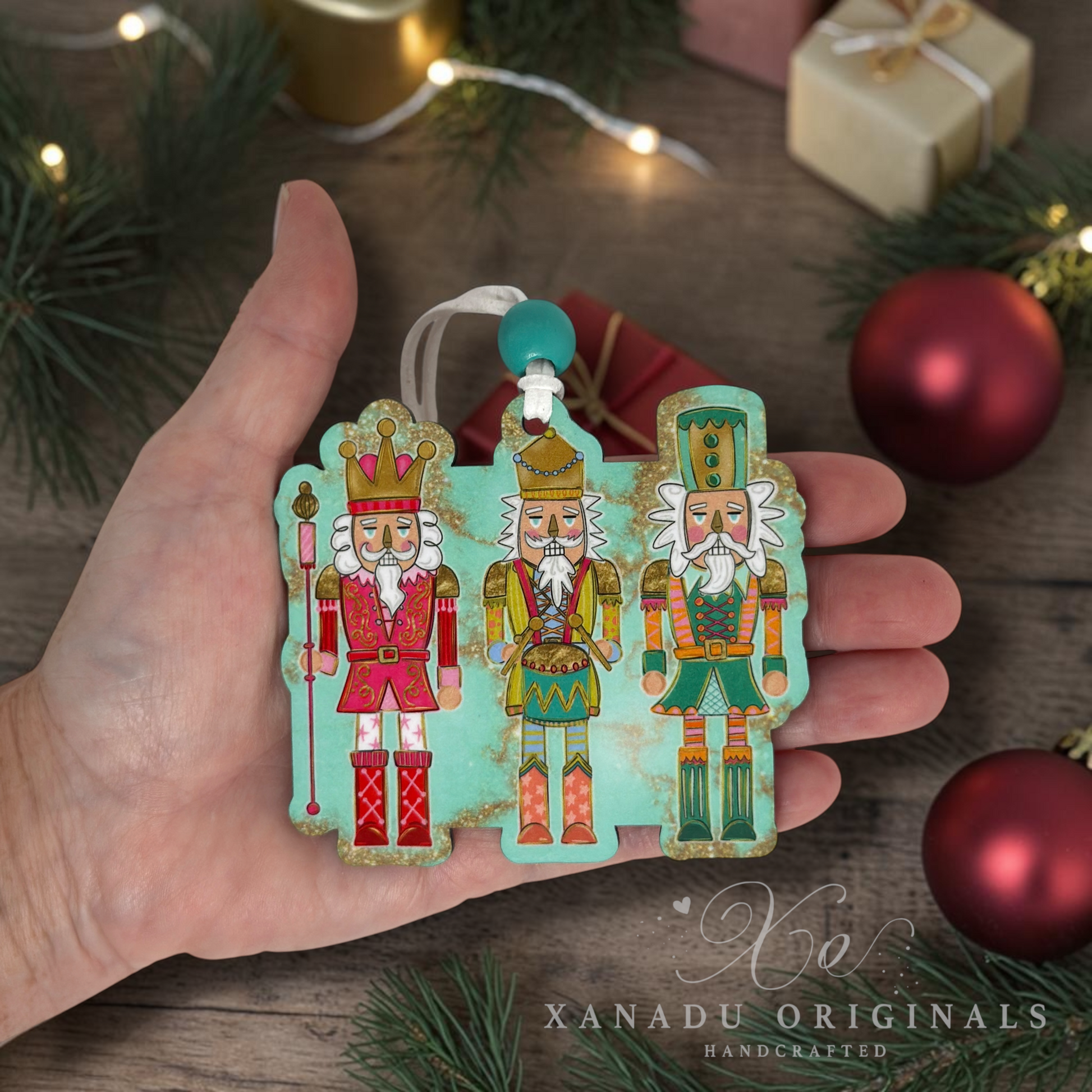 A hand holds a turquoise nutcracker ornament featuring three festive nutcracker figures with gold, red, and green details. The background is a rustic wooden surface decorated with pine branches, fairy lights, and red ornaments.