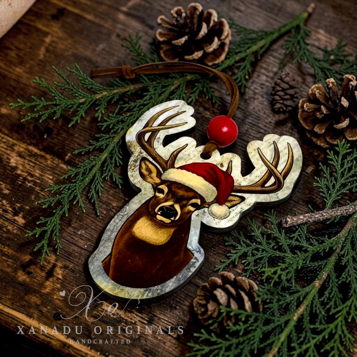 A rustic deer head wood ornament with a newsprint background design lying flat on a dark wooden surface surrounded by evergreen branches and pine cones.

