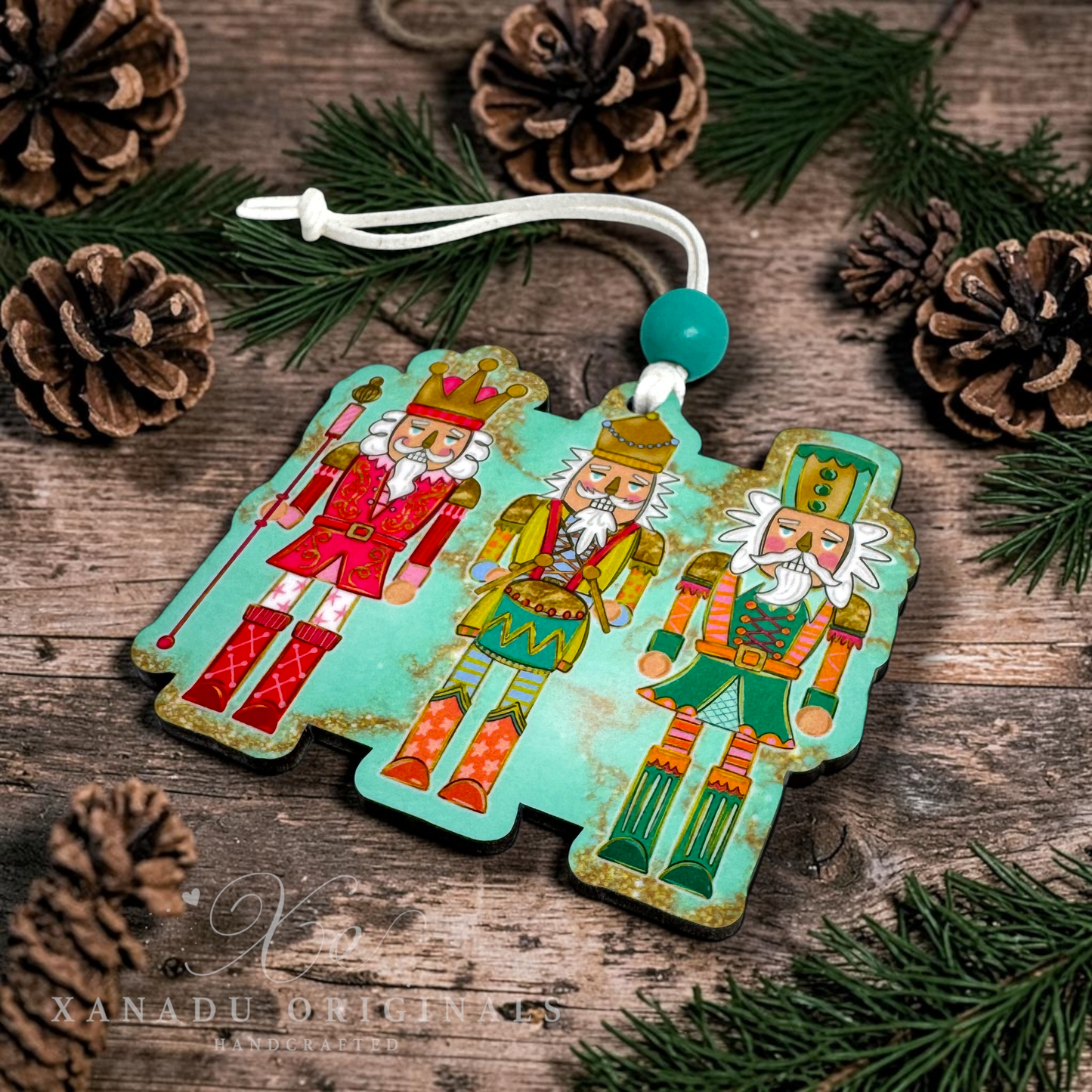 A three-piece Christmas nutcracker ornament set with a distressed turquoise background and colorful illustrations of three nutcrackers, displayed on a rustic wooden surface with pine needles and pinecones. A white hanging string with a turquoise bead is attached to the top.